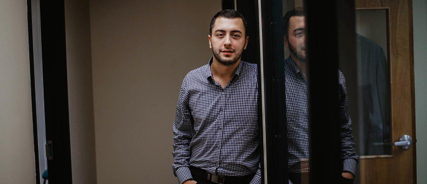 A young man looking at the camera, leaning against the side of an open doorway.